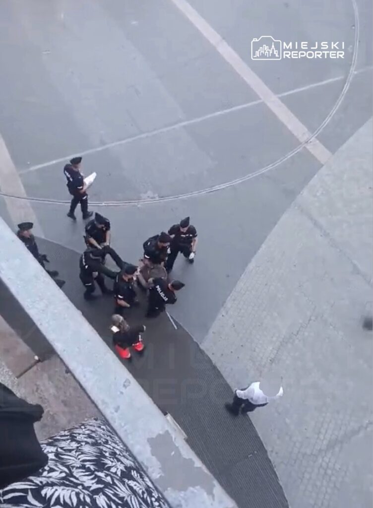 Grupa policjantów w mundurach zatrzymuje osobę na placu, w otoczeniu innych przechodniów.