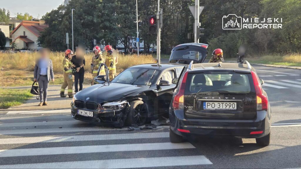 Czarny samochód osobowy z uszkodzeniami stoi na pasach, obok grupka strażaków oraz świadek sytuacji w garniturze.