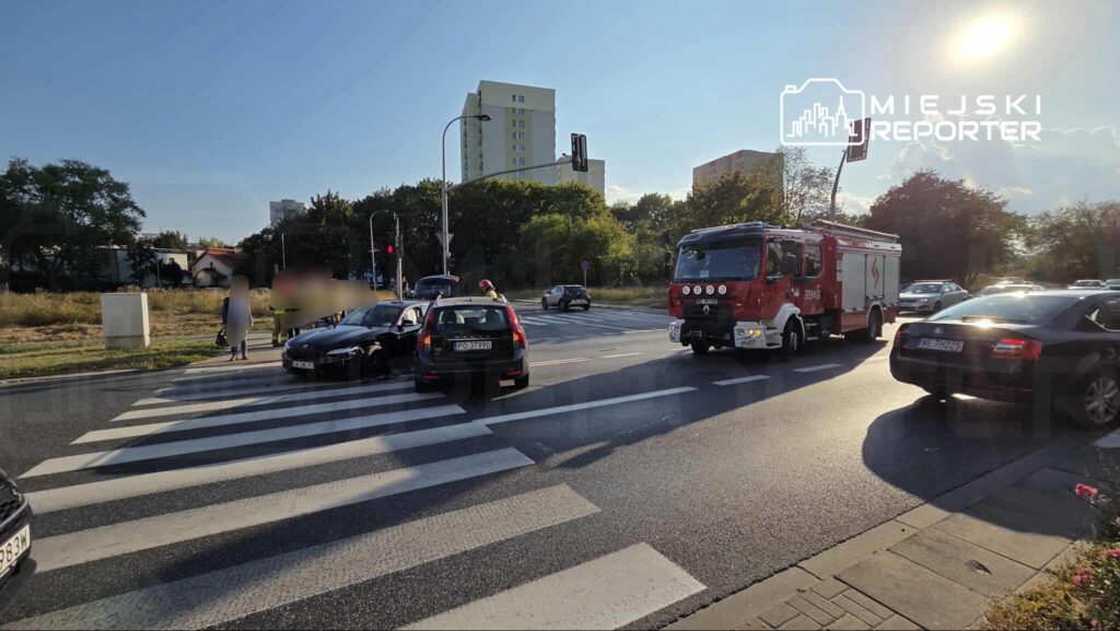 Na przejściu dla pieszych zatrzymane są dwa samochody, a obok stoi straż pożarna. W tle widać budynki i zielone drzewa.
