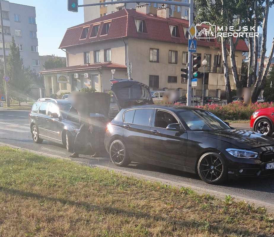Mężczyzna przy samochodzie z otwartym bagażnikiem, stojący obok drugiego auta na ruchliwej ulicy.