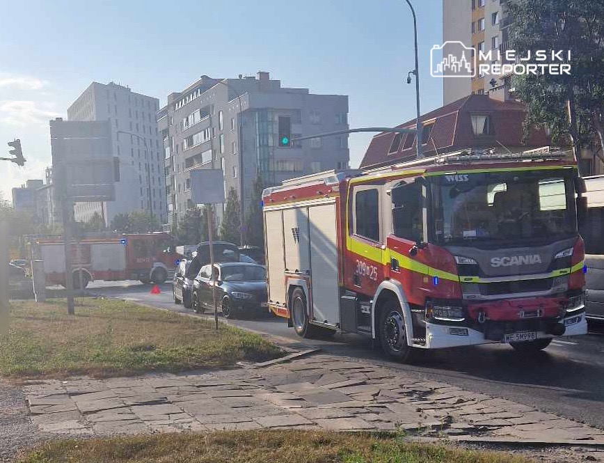 Wozidło strażackie zatrzymane na skrzyżowaniu obok nowoczesnych budynków, z włączonymi sygnałami świetlnymi.