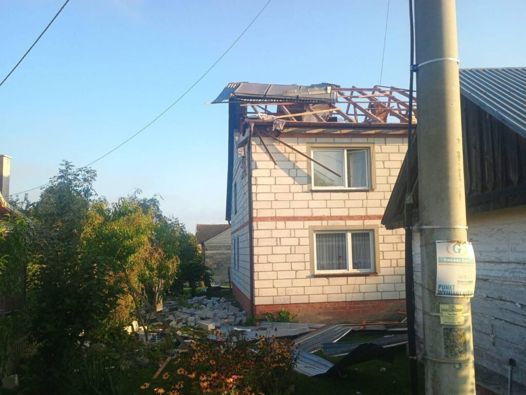 Zniszczony dach na budynku mieszkalnym, wokół leżące fragmenty materiałów budowlanych i zniszczona roślinność.