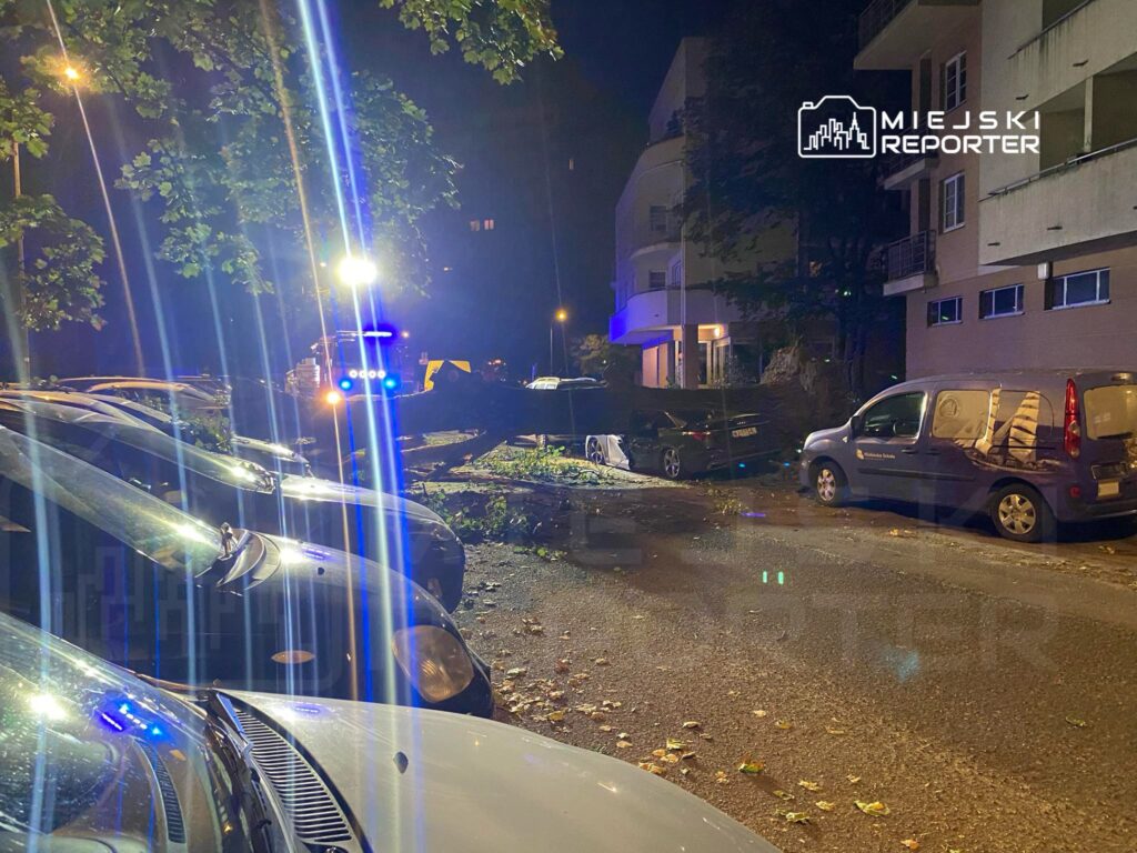 Połamane drzewo leży na drodze obok zaparkowanych samochodów, a w tle widoczne są pojazdy służb ratunkowych z włączonymi światłami.