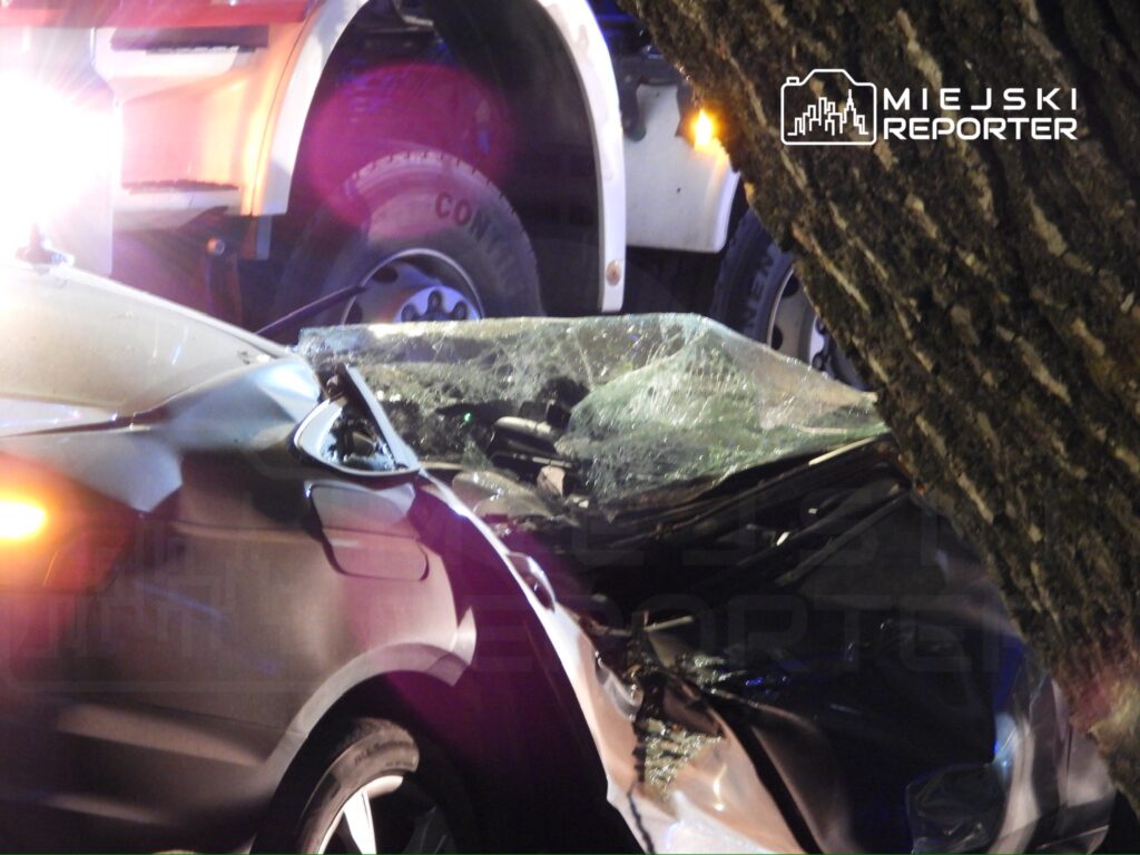Uszkodzone auto osobowe z rozbitą szybą, stojące obok dużego pojazdu, w tle widoczne migające światła policyjne.