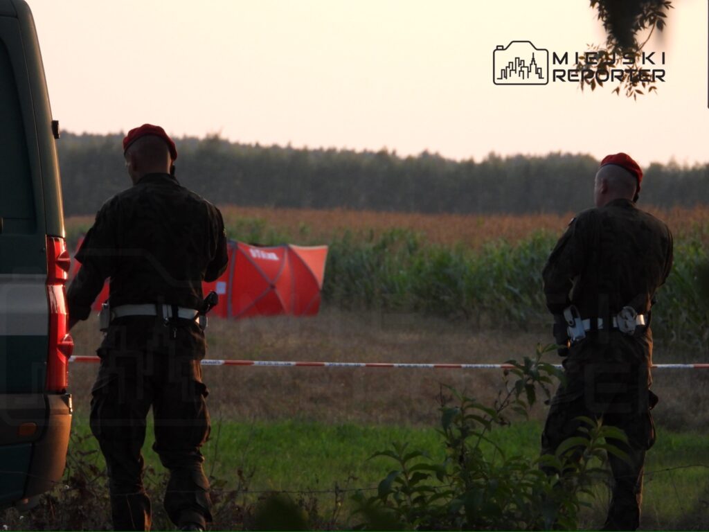 Dwaj żołnierze w mundurach z czerwonymi beretami obserwują teren przy zielonym pojeździe, w tle widoczny namiot.