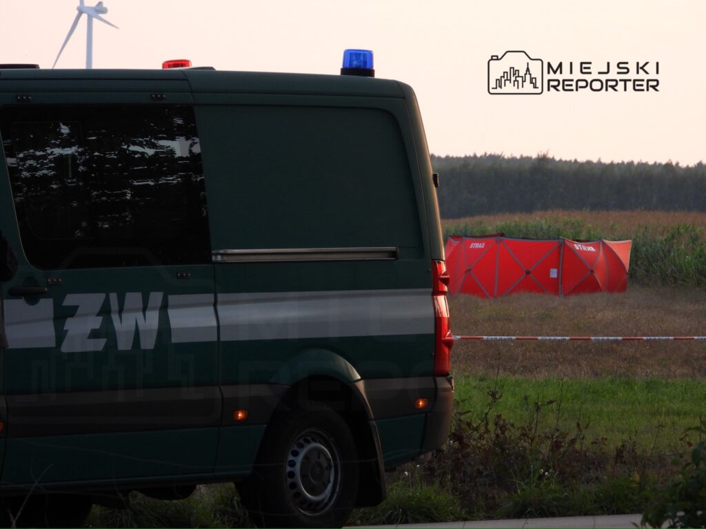 Zielony pojazd służbowy z niebieskimi i czerwonymi sygnalizatorami stoi obok czerwonego namiotu ratunkowego na polu.