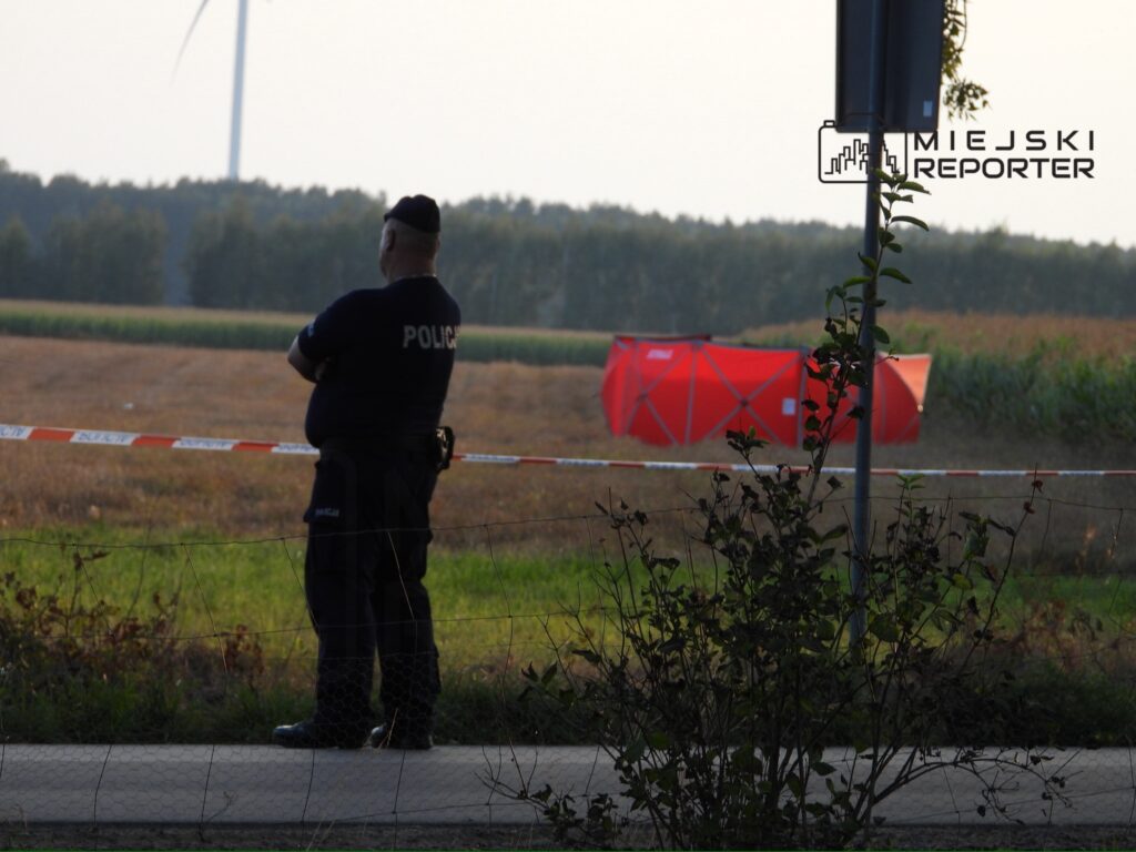 Policjant w mundurze stoi przy ogrodzeniu w pobliżu pola, gdzie widoczny jest czerwony namiot.