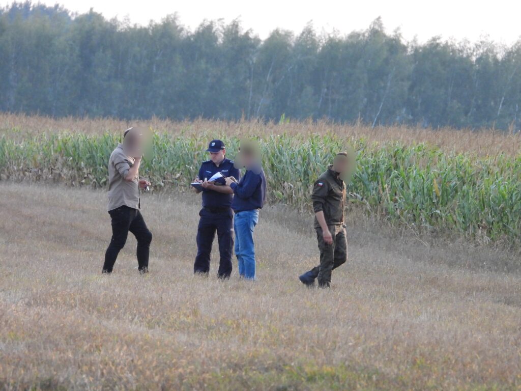 Czterej mężczyźni rozmawiają na polu, w tle rośnie kukurydza, jeden z nich nosi mundur policyjny.