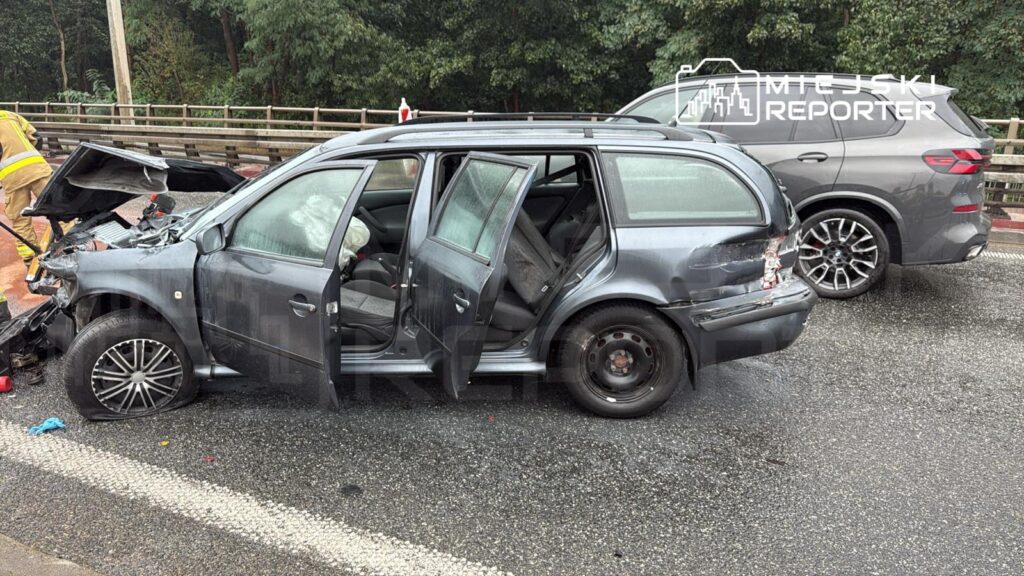 W wyniku wypadku drogowego uszkodzone auto osobowe z otwartymi drzwiami stoi na jezdni, obok pracują strażacy.