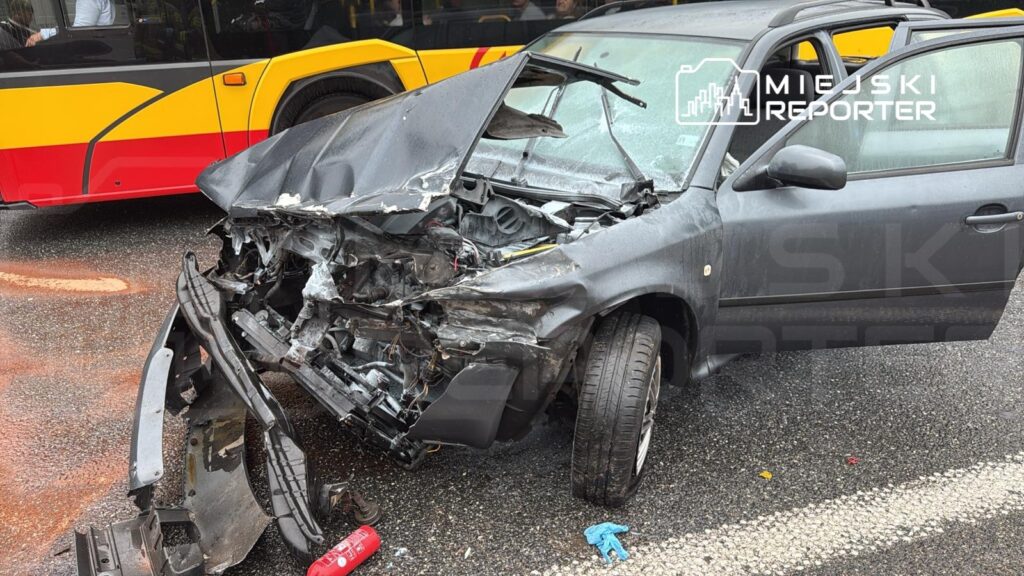 Wgniecione auto osobowe leży na drodze, obok niego widoczny czerwony gaśnica oraz niebieska rękawiczka. W tle przejeżdżający autobus.