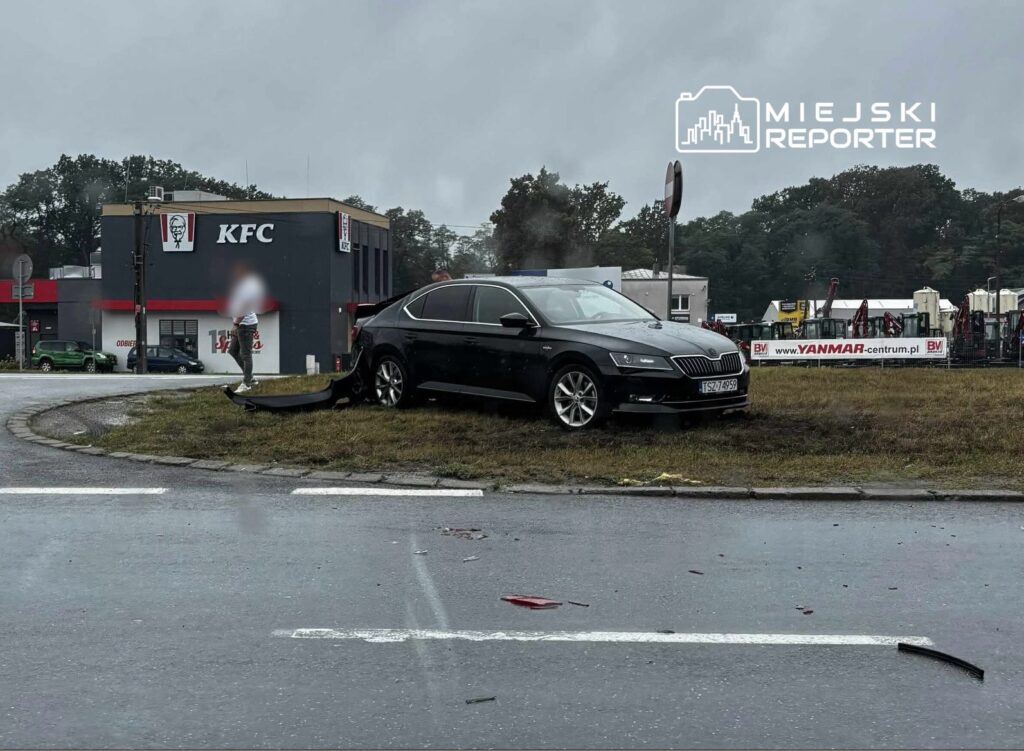 Czarny samochód osobowy stoi na trawie obok drogi, a obok niego mężczyzna w białej koszulce bada uszkodzenia pojazdu.