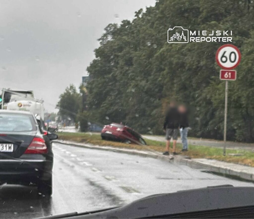 Czerwony samochód leży na poboczu drogi, obok dwóch mężczyzn rozmawiających w deszczowej aurze. W tle widoczny znak ograniczenia prędkości.
