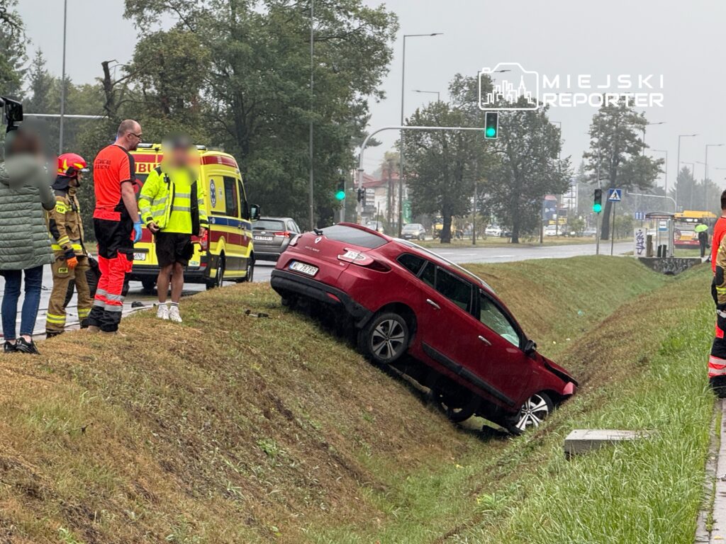 Czerwony samochód osobowy w rowie, obok stoją strażacy i ratownicy medyczni w odblaskowych kamizelkach.