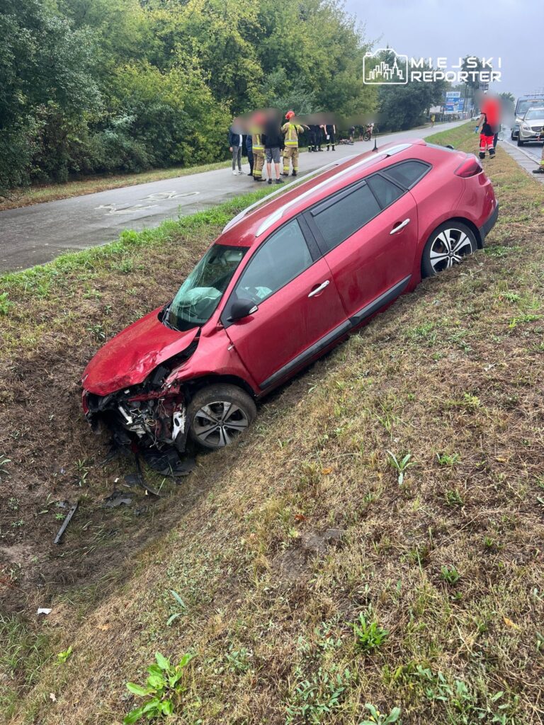 Czerwony samochód osobowy w rowie, obok grupy osób w mundurach strażackich i ratowników medycznych.