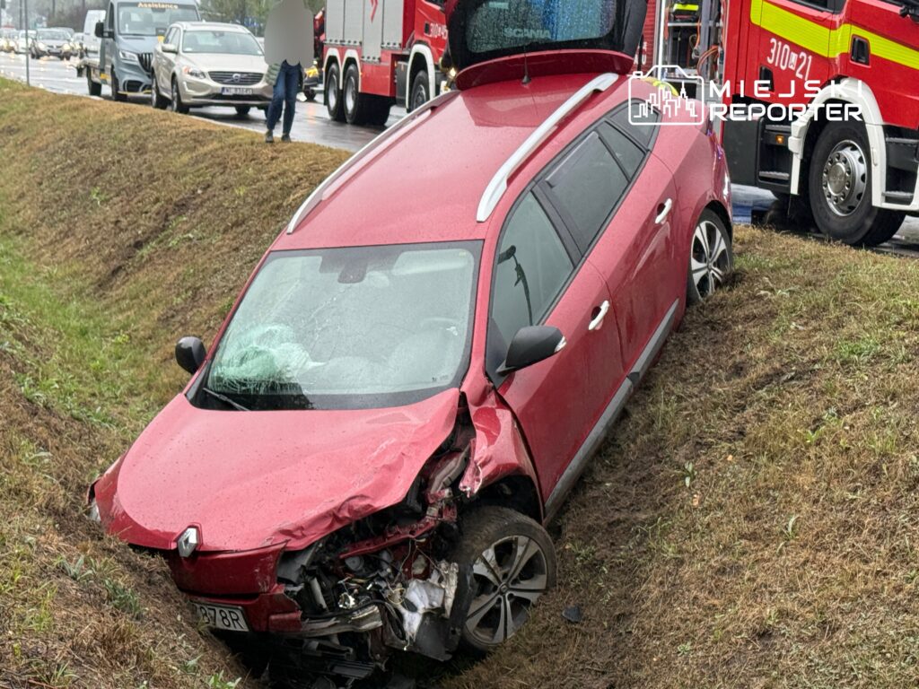 Czerwony samochód osobowy w rowie, z uszkodzoną przednią częścią, obok stojące pojazdy i wzywający pomoc strażak.
