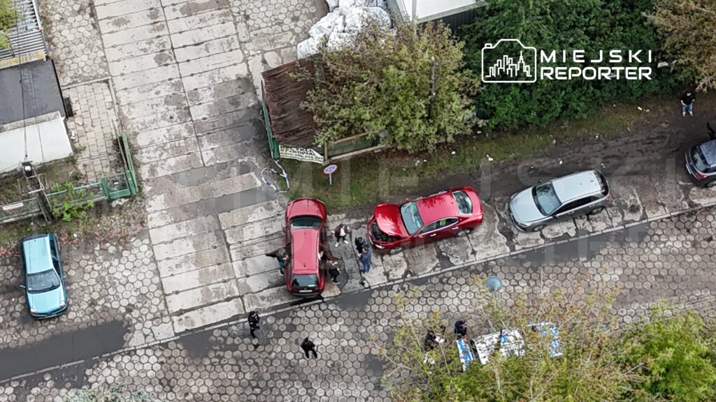 Na ulicy stoją dwa czerwone samochody, przy których grupka osób rozmawia, a obok widać niebieski pojazd.
