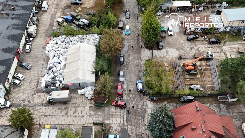 Widok z góry na plac z samochodami, w tym czerwonym pojazdem, oraz stosy plastikowych worków obok białego namiotu. Policjanci prowadzą czynności na drodze.