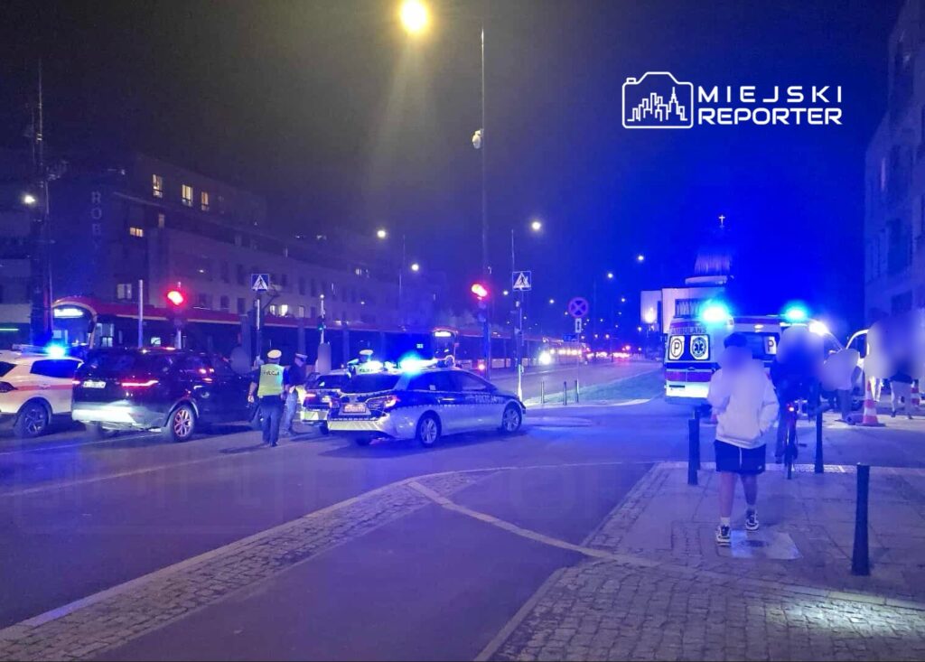 Policjanci kierują ruchem na skrzyżowaniu, obok zatrzymane pojazdy i karetka, w tle nocne miasto z oświetleniem.