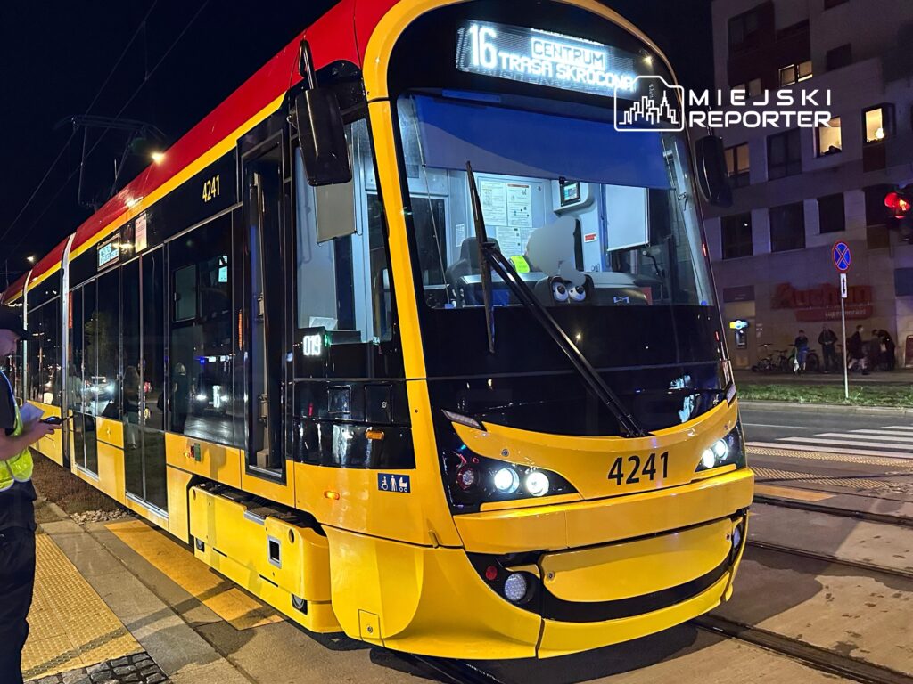 Tramwaj linii 16 zatrzymany na przystanku w nocy, obok mężczyzna w kamizelce odblaskowej sprawdzający telefon.