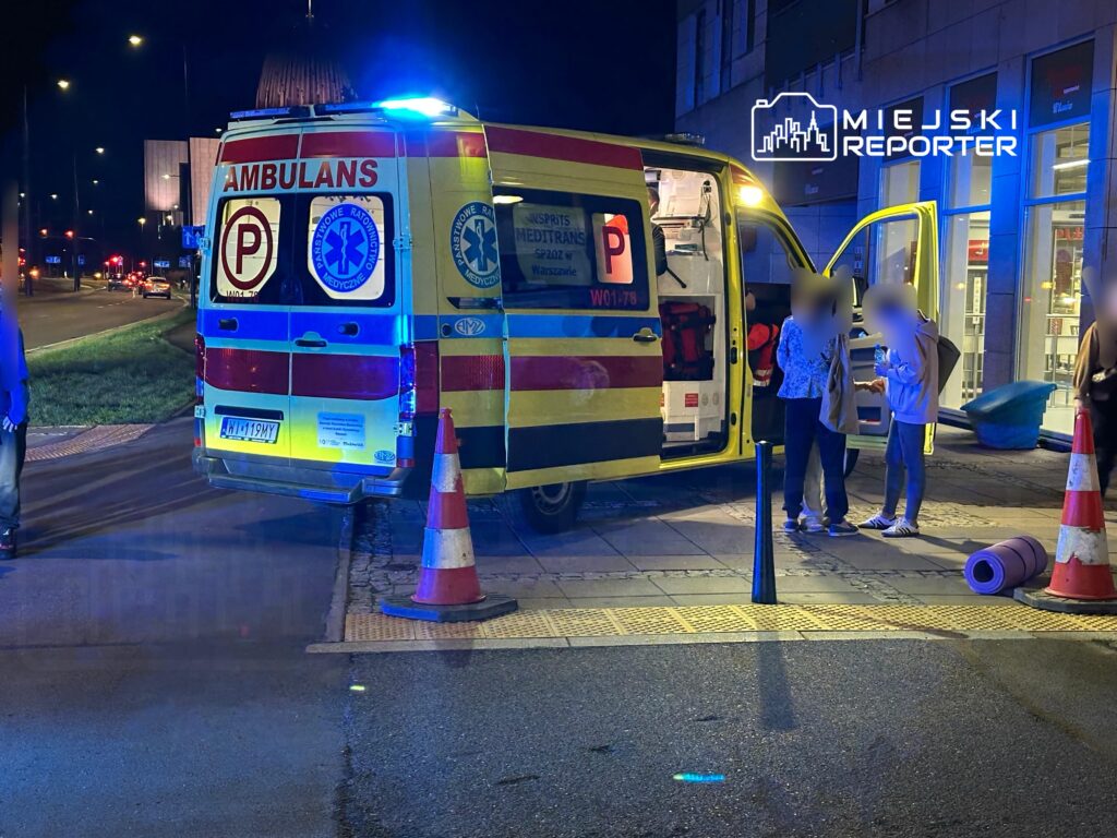 Ambulans z włączonymi światłami stoi na chodniku, obok dwóch osób rozmawiających z ratownikami medycznymi.