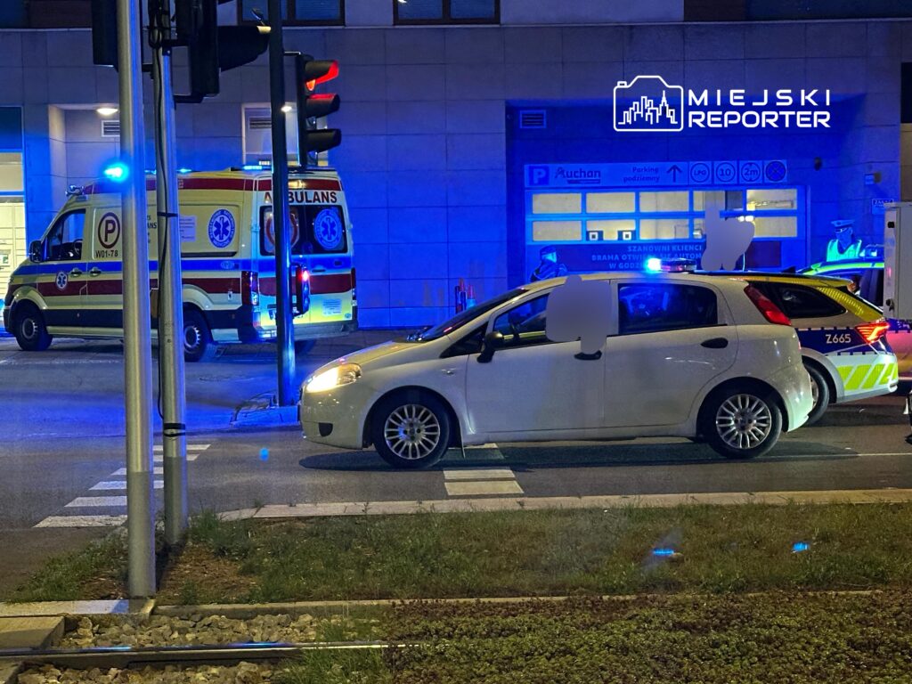 Ambulans z włączonymi sygnałami świetlnymi zatrzymany obok białego samochodu osobowego na ulicy w nocy.