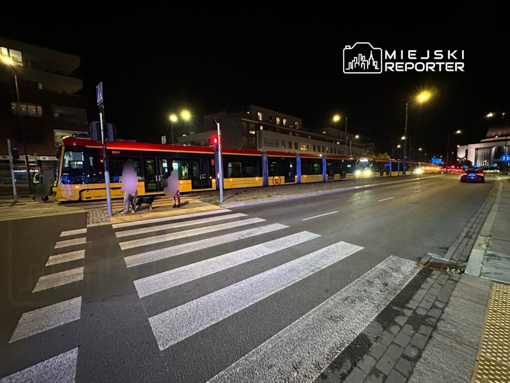 Tramwaj z numerem 4241 zatrzymany na przystanku, obok przejścia dla pieszych w oświetlonej nocą ulicy.