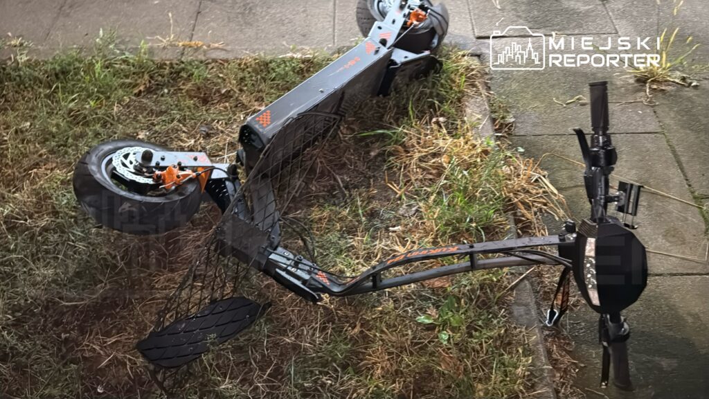 Złamana hulajnoga elektryczna leży na trawie obok chodnika, z widocznymi uszkodzeniami.