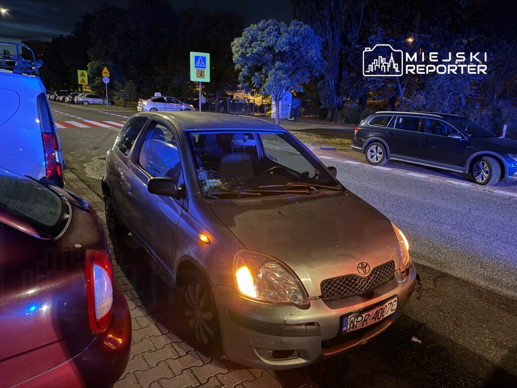 Zniszczony samochód osobowy zaparkowany na chodniku w nocy, w tle widoczne znaki drogowe i inne pojazdy.