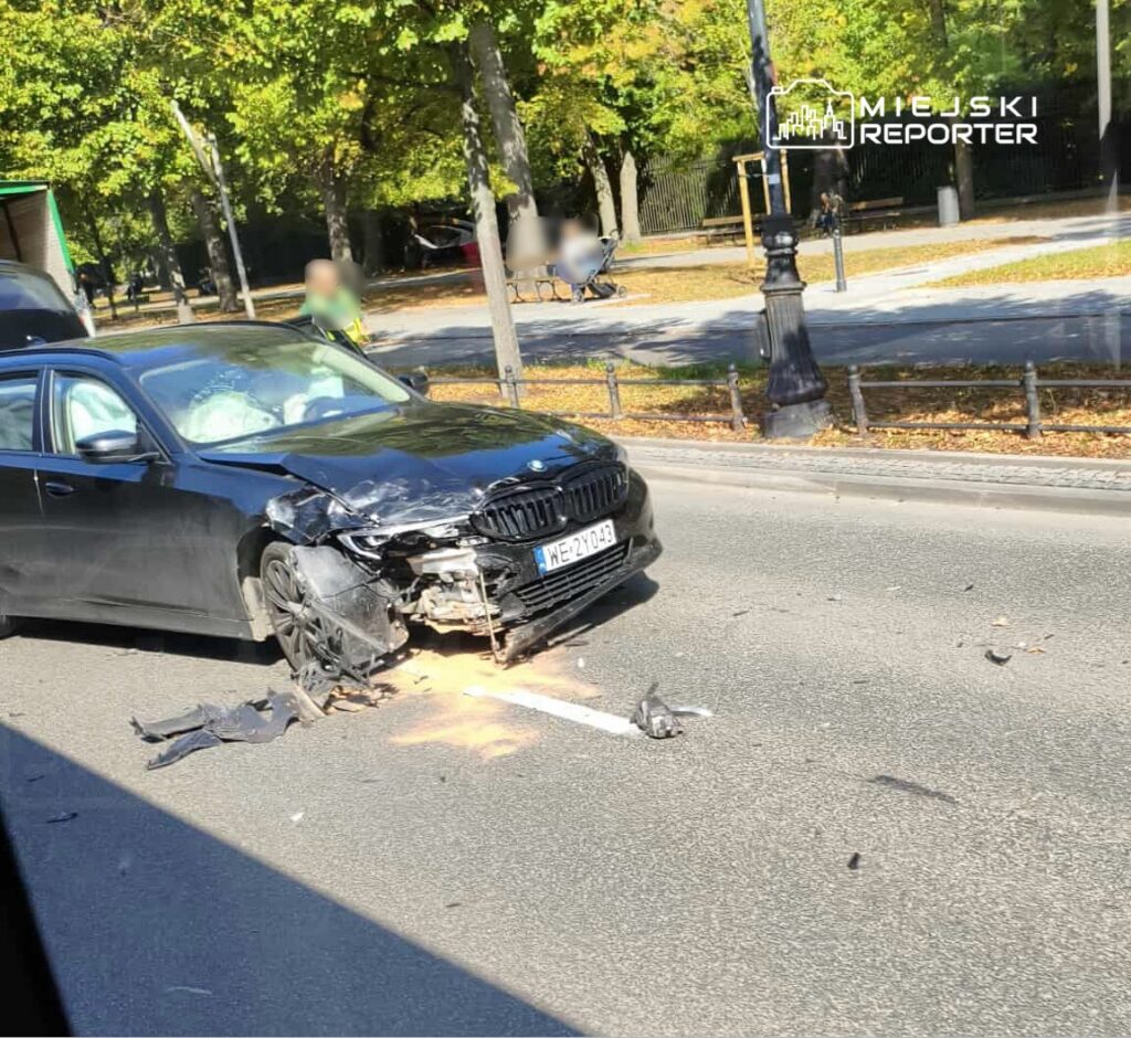 Czarny samochód osobowy z uszkodzoną przednią częścią stoi na jezdni, obok widoczne zniszczenia i kawałki karoserii.