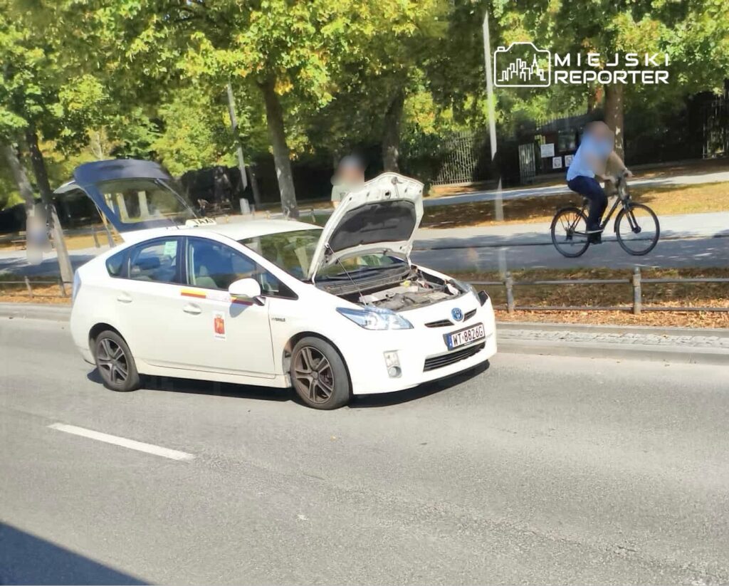Biały samochód osobowy z otwartą maską stoi na drodze, obok przejeżdża rowerzysta. W tle widać drzewa i chodnik.
