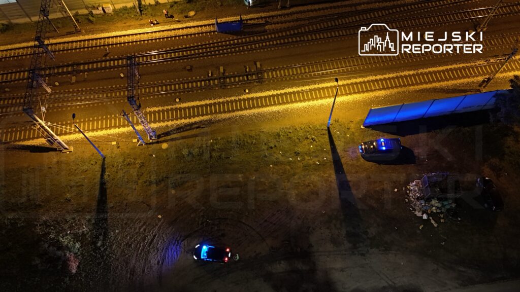 Dwa radiowozy policyjne zaparkowane w pobliżu torów kolejowych oświetlonych nocnymi lampami.
