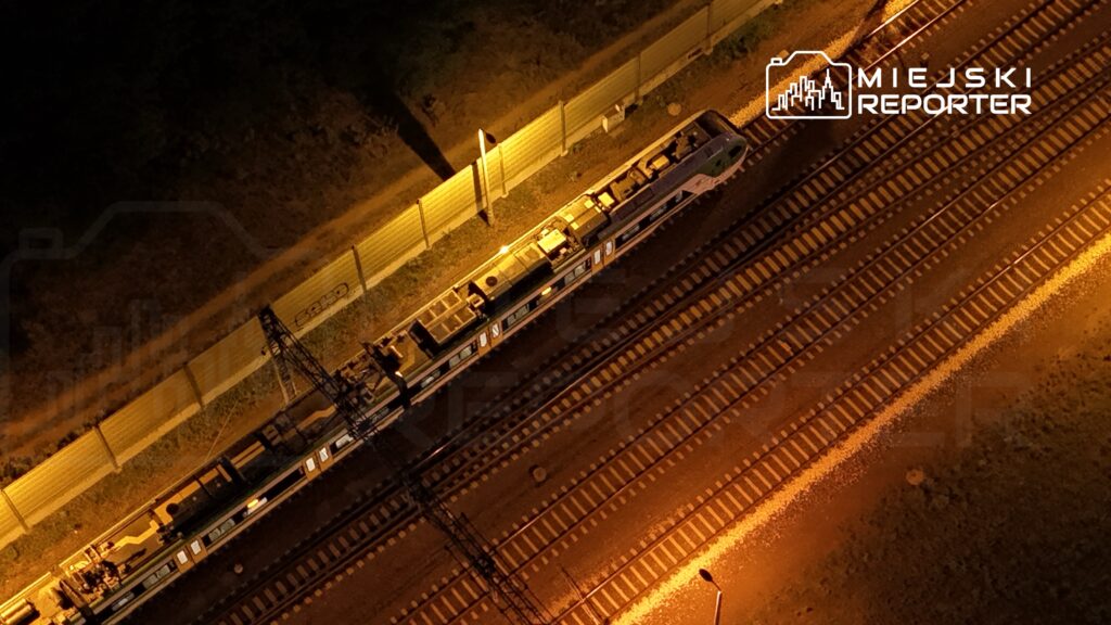 Pociąg stojący na torach, oświetlony lampami w nocy, otoczony metalowym płotem i zielenią.
