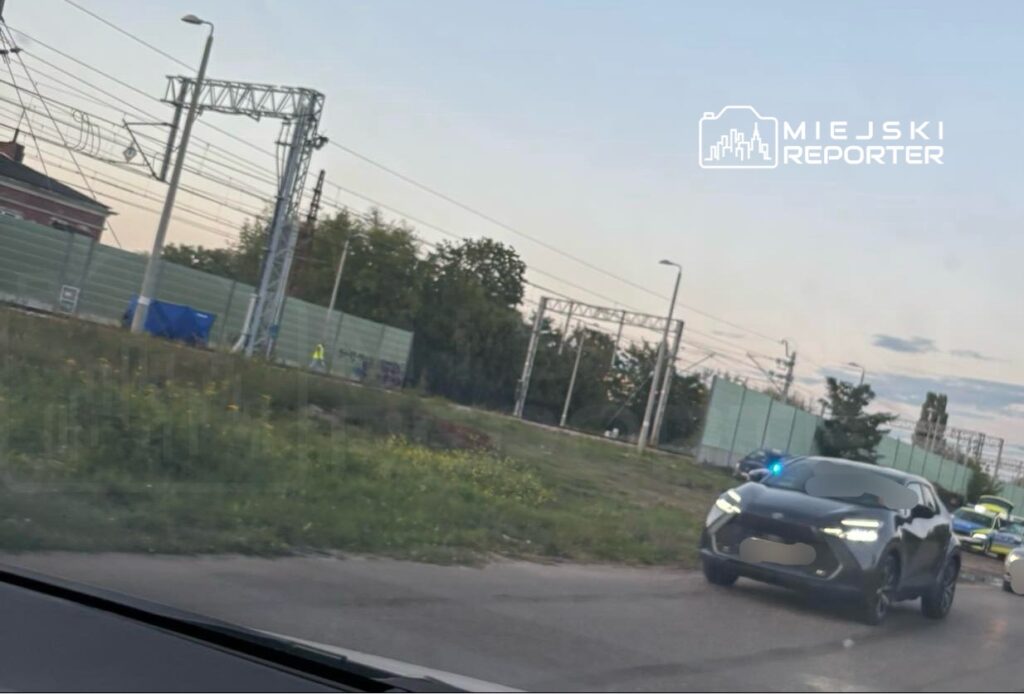 Policyjny samochód z włączonymi sygnałami świetlnymi przemieszcza się wzdłuż drogi obok torów kolejowych.