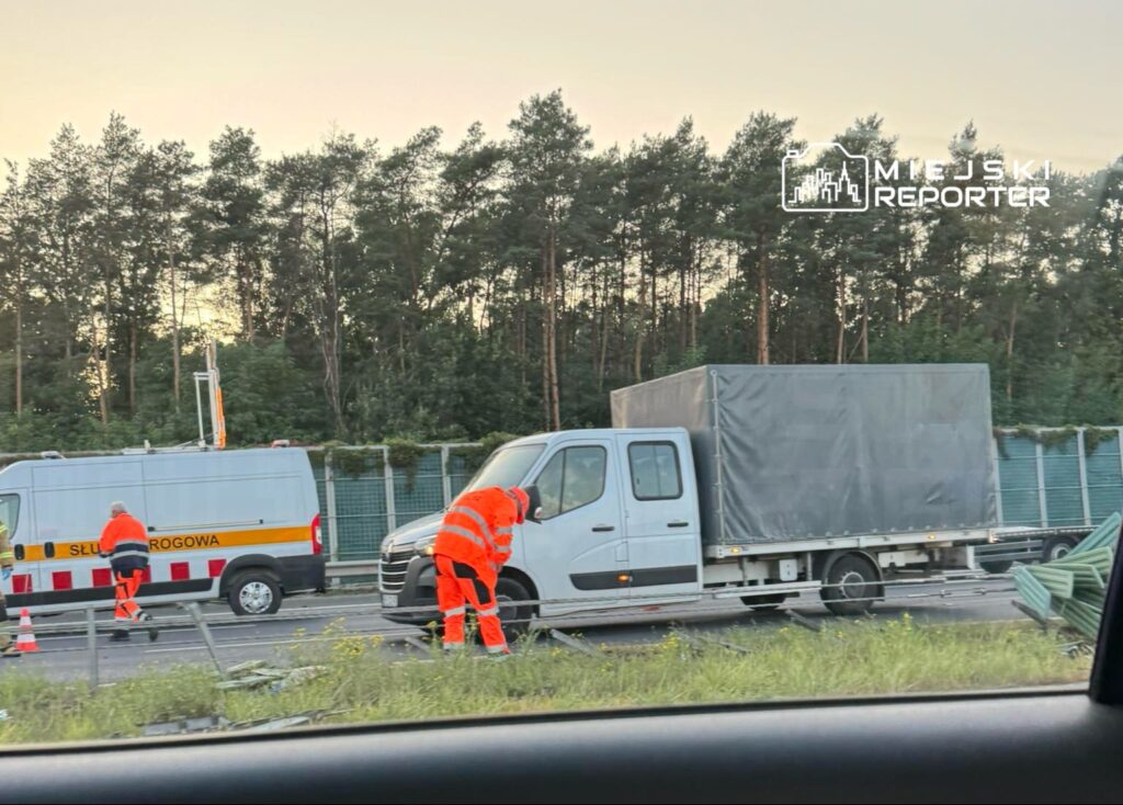 Dwaj pracownicy w pomarańczowych uniformach naprawiają uszkodzone barierki na drodze, obok zaparkowanego pojazdu.