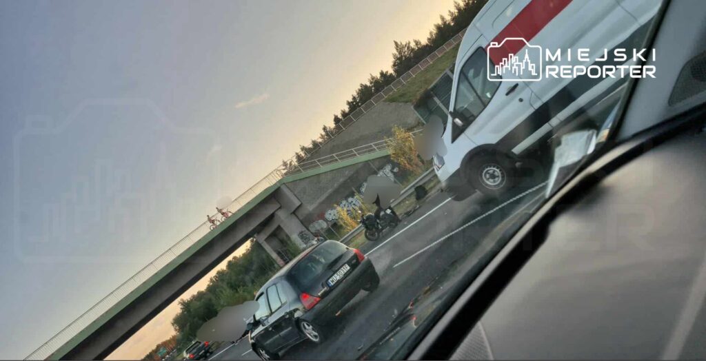 Ruch samochodowy na drodze, z motocyklistą zatrzymanym obok graffiti na murze, w tle widoczny most.