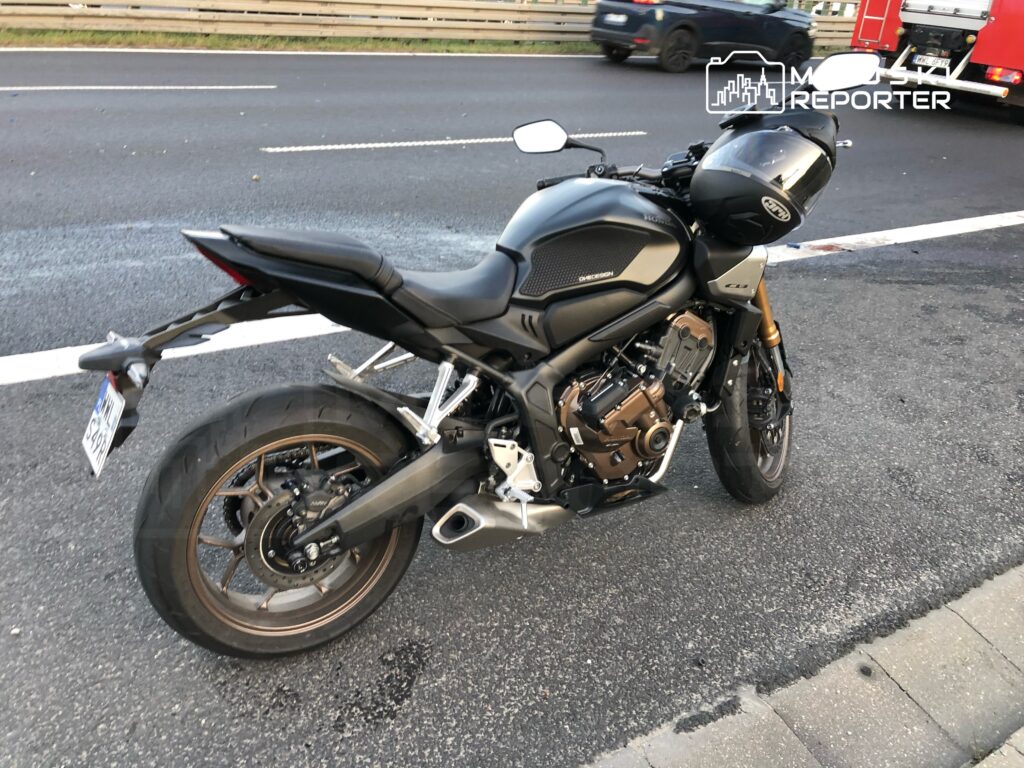 Motocykl w kolorze czarnym z widocznym silnikiem, zaparkowany na poboczu drogi obok ruchliwej jezdni.