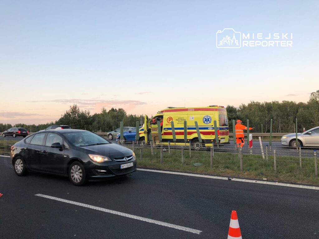 Karetka pogotowia zatrzymana na drodze, a pracownik w pomarańczowym uniformie kieruje ruchem w pobliżu.