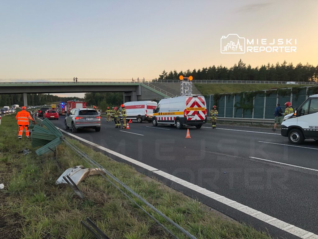 Zespół ratunkowy oraz pojazdy służb drogowych pracują na miejscu wypadku na autostradzie, w otoczeniu stojących samochodów.