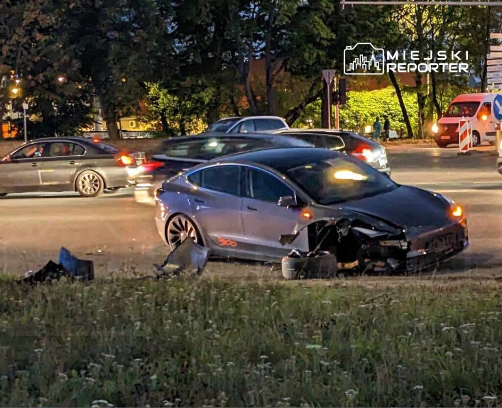 Zniszczony samochód osobowy stoi na poboczu drogi, obok przejeżdżające pojazdy w ruchu nocnym.