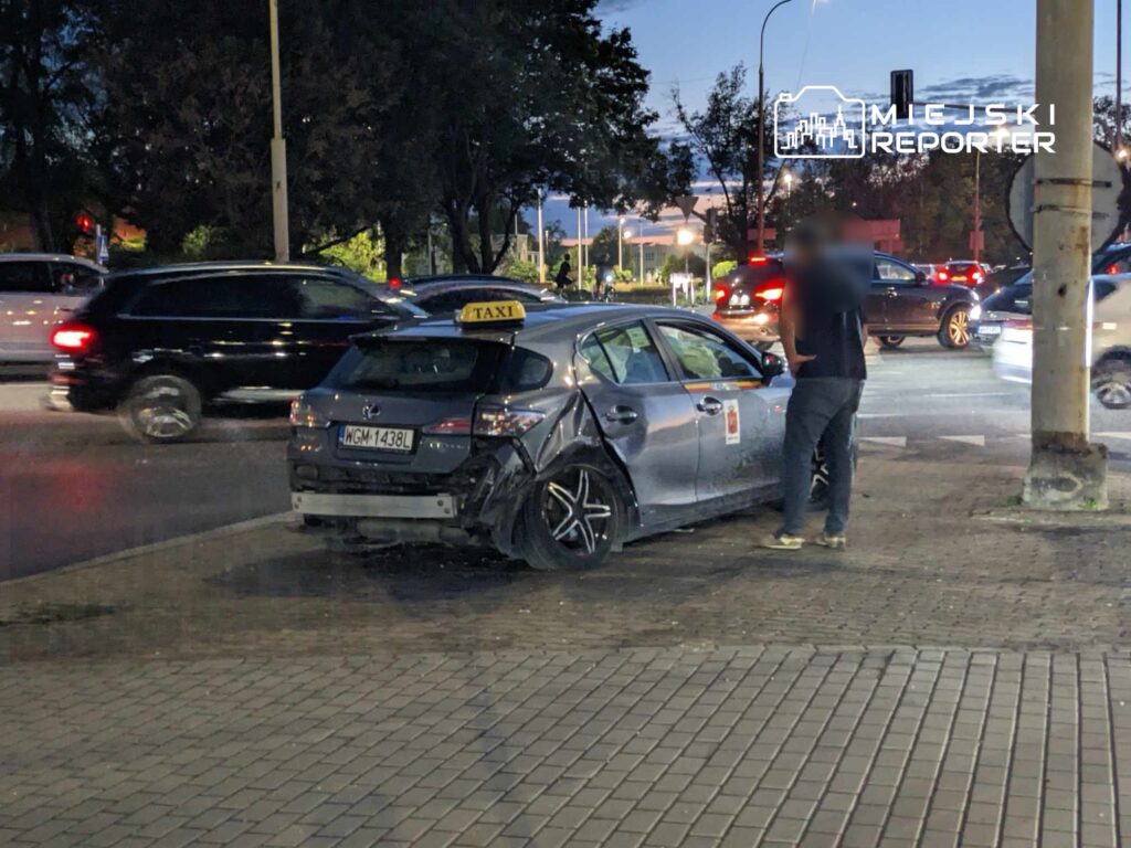 Uszkodzone taxi stoi na chodniku, obok kierowca w czarnej koszulce rozmawia z inną osobą, w tle ruch uliczny.