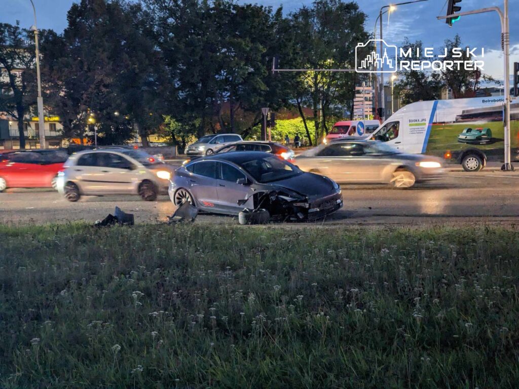 Czarny samochód osobowy z uszkodzoną przednią częścią stoi na poboczu, obok ruchliwej drogi w godzinach wieczornych.