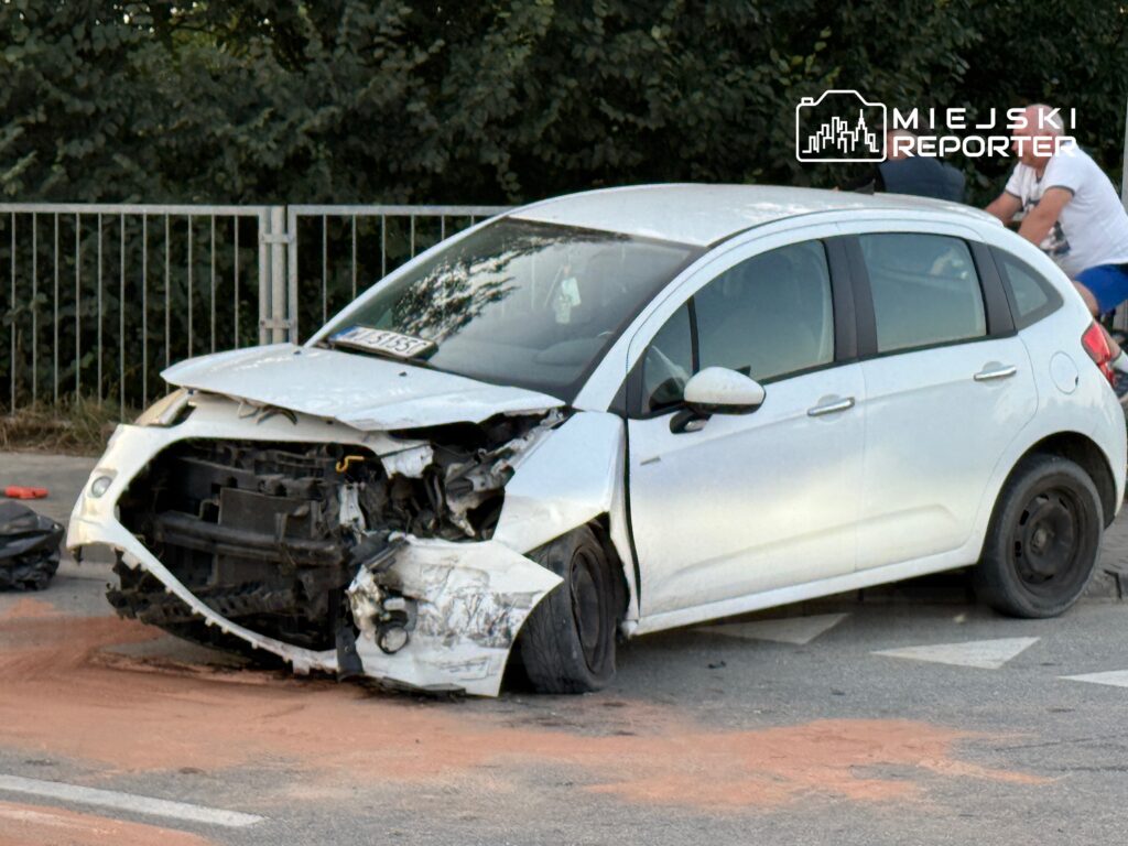 Biały samochód osobowy z uszkodzeniami przodem stoi na drodze, obok dwóch mężczyzn w tle.