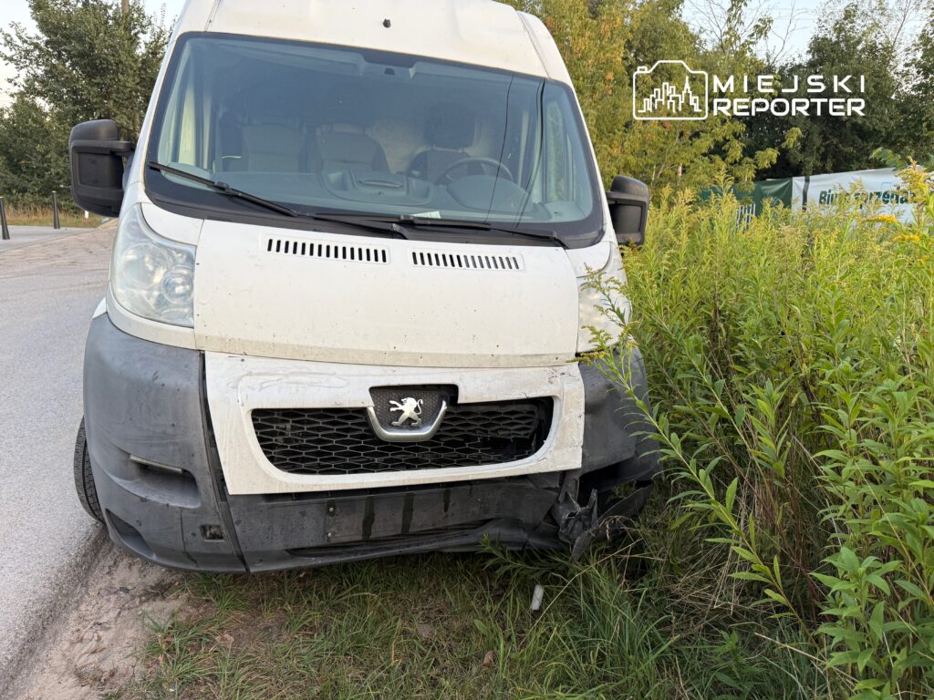 Uszkodzony biały samochód dostawczy zaparkowany na poboczu drogi, otoczony wysoką trawą i krzewami.