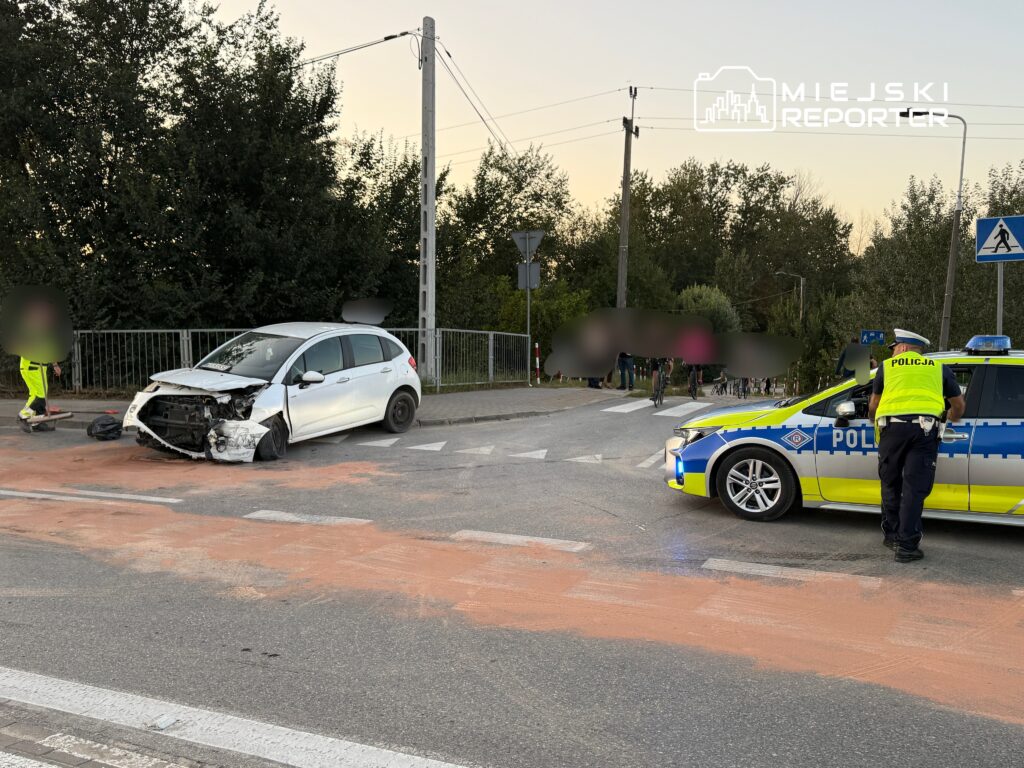 Uszkodzony samochód osobowy stoi na skrzyżowaniu, obok policyjny radiowóz, a w tle grupka pieszych.