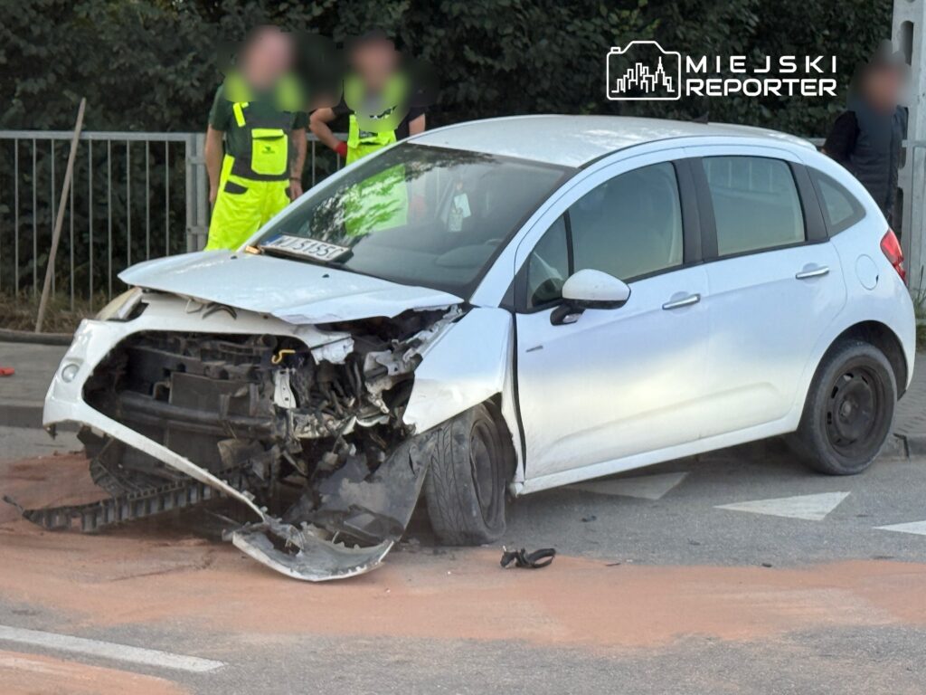 Uszkodzony biały samochód osobowy stoi na drodze, obok grupy osób w odzieży roboczej.