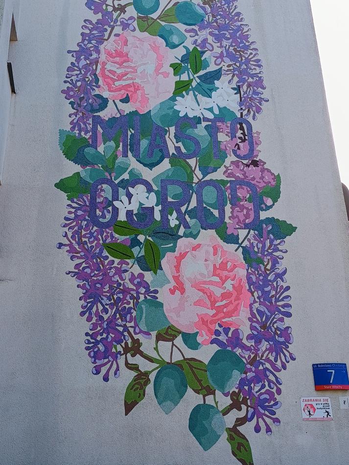 Na ścianie budynku znajduje się kolorowy mural z kwiatami i napisem "MIASTO OGRÓD".