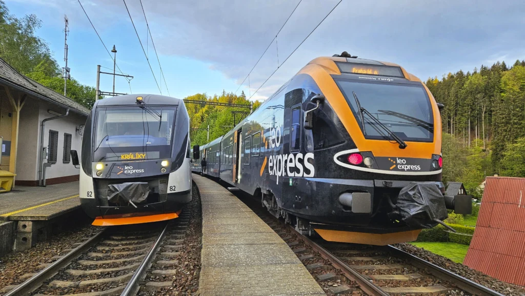 Pociągi Leo Express mijają się na stacji, otoczone zielenią drzew i budynkami w tle.