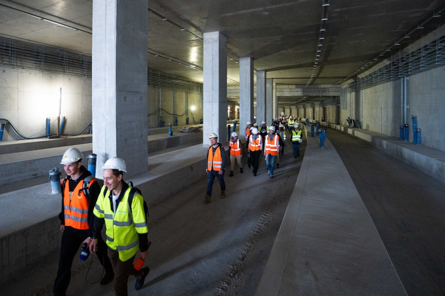 Grupa pracowników w kaskach i kamizelkach odblaskowych spaceruje po nowym tunelu budowlanym.