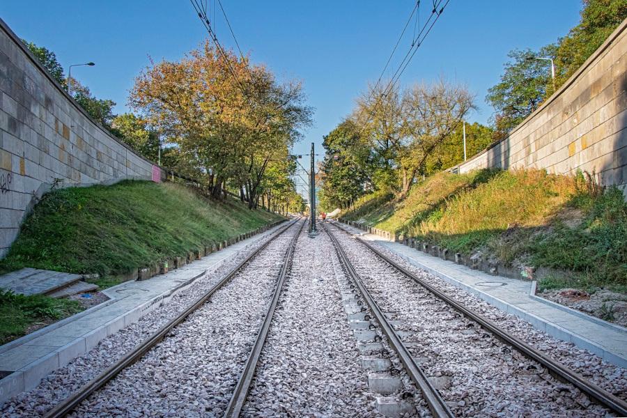 Tory tramwajowe biegnące przez zielony teren, otoczone murami i drzewami w słoneczny dzień.