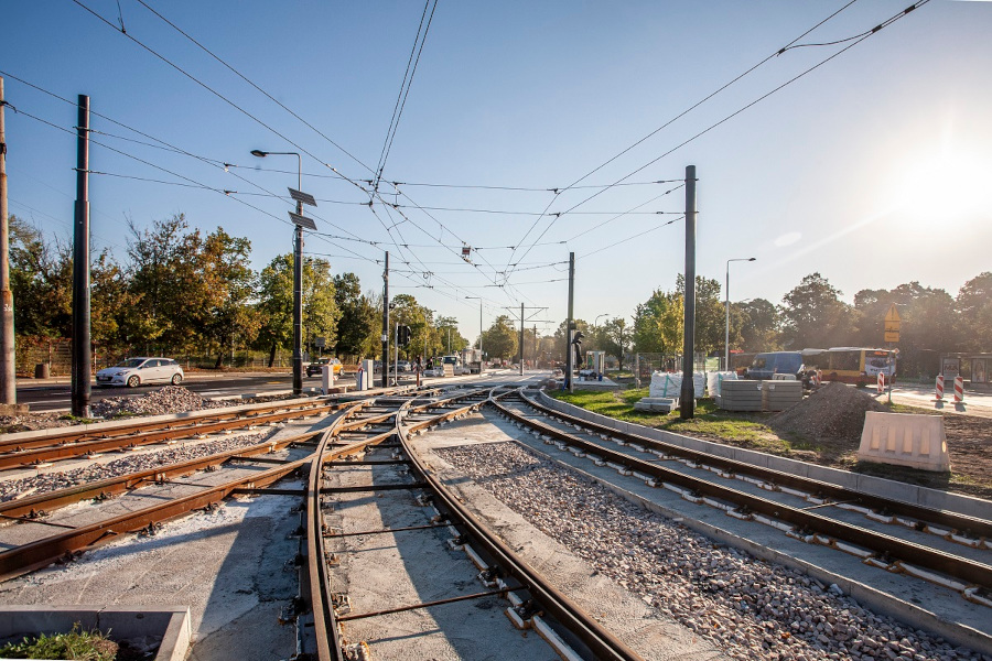 Torowisko tramwajowe skręcające w prawo w nowo budowanej infrastrukturze miejskiej, w tle widoczna zieleń i pojazdy.